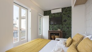 A hotel room with a bed, a TV mounted on a green leaf-patterned wall, a wooden side table, and a view of the city through a large window. - S. Bento Downtown Apt (Oporto)