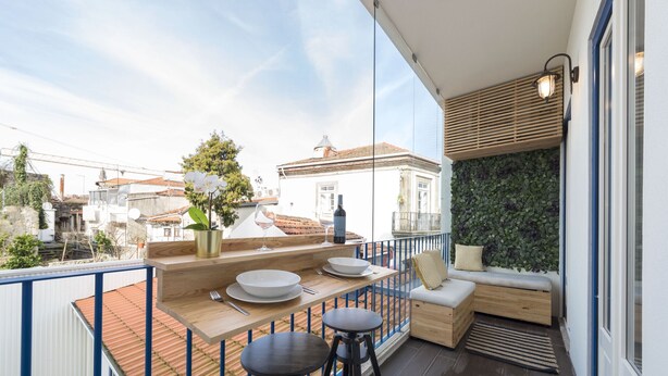 A balcony with a wooden table, two stools, a cushioned bench, and a wall-mounted light fixture. - S. Bento Downtown Apt (Oporto)