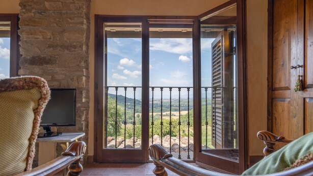 A room with a stone wall, a television, a patterned rug, and a view of a balcony with a railing. - La Terrazza by Interhome (Vicchio)