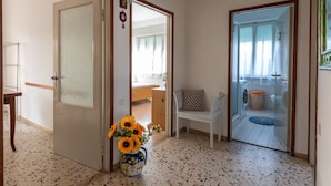 A hallway with a door leading to a bedroom, a small seating area with a chair, and a washing machine in the adjacent room. - Apartment Angela by Interhome (Follonica)