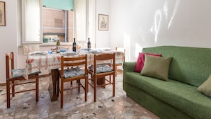 A dining area with a table set for four, surrounded by wooden chairs with patterned cushions. A green sofa with a red pillow is against the wall. - Apartment Angela by Interhome (Follonica)