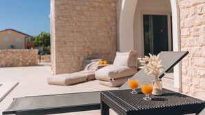 A sunlit outdoor seating area with a stone wall, a lounge chair, and a table with two glasses of orange juice.