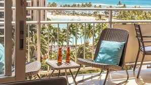Uma varanda com cadeiras de vime, uma mesa pequena com garrafas e vista para a praia e o oceano.
