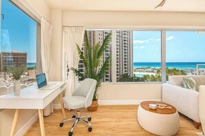 A modern room with a desk, chair, round ottoman, and large windows offering a view of the ocean and high-rise buildings. - 2BR/2BA Waikiki Ocean View Condo w/ 2 Balconies (Honolulu)