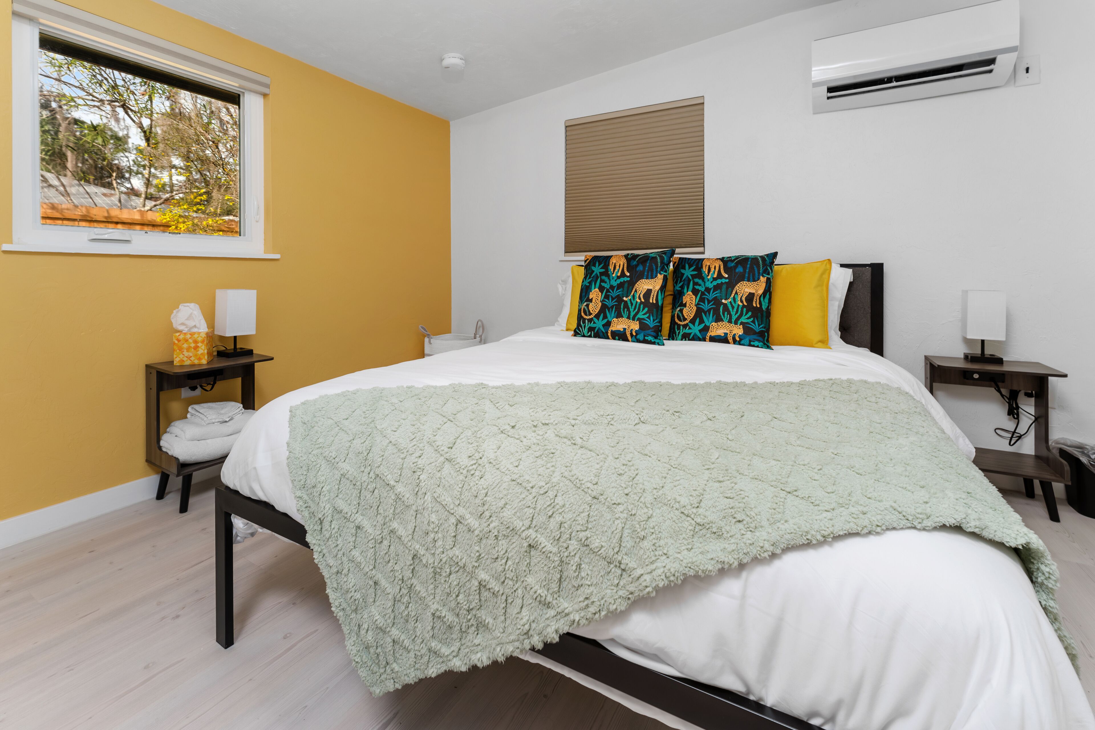 A bedroom with a bed, a window, an air conditioner, and a bedside table with a lamp.
