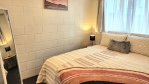 A bedroom with a bed, a nightstand, a lamp, a window with curtains, a painting on the wall, and a mirror. - CQN's (Wellington)
