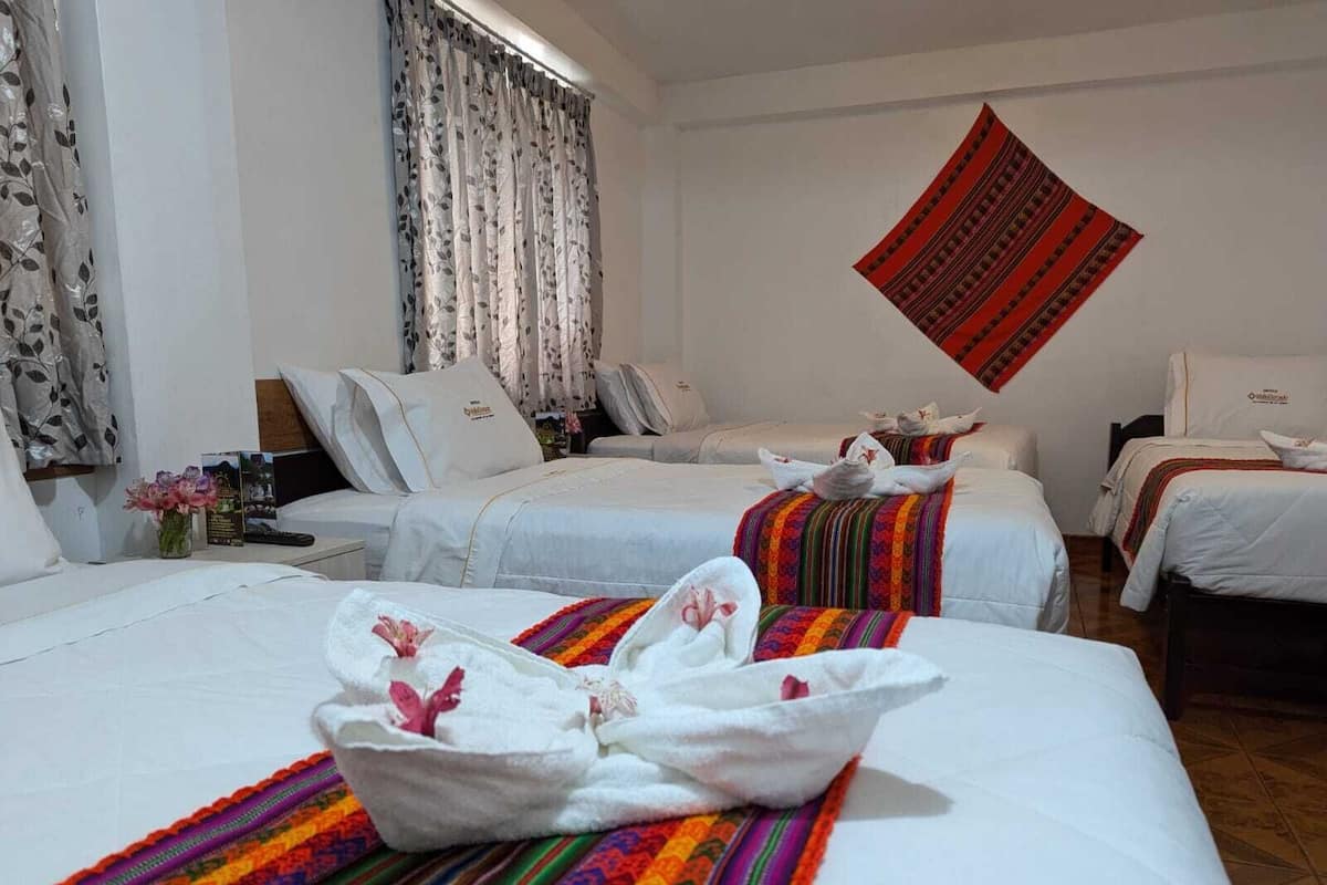 A hotel room with two beds, a large window with curtains, a wall hanging, and a small table with a vase of flowers.