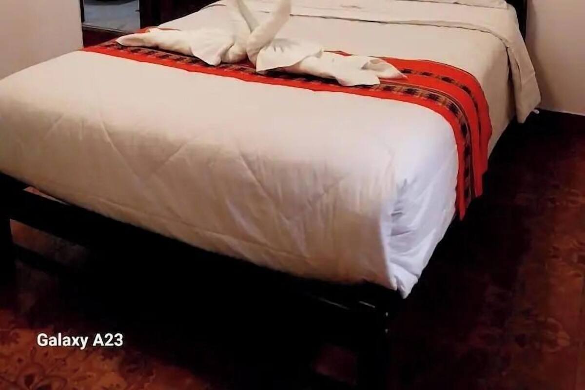 A neatly made bed with a wooden headboard, white bedding, and a red and black patterned blanket.