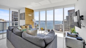 Una sala de estar moderna con un sofá grande de color gris, una mesita auxiliar y vistas al mar y a edificios a través de ventanales.
