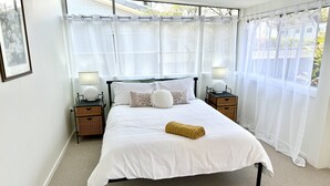 Una cama bien hecha con sábanas blancas, dos mesitas de noche con lámparas y cortinas translúcidas en una ventana.