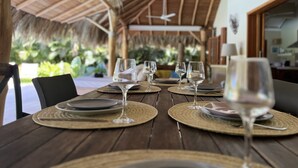 A dining table set with plates, cutlery, and wine glasses, under a thatched roof with a view of greenery. - Las Terrenas - Caribbean Villa for 6 People - Exceptional Location (Las Terrenas)