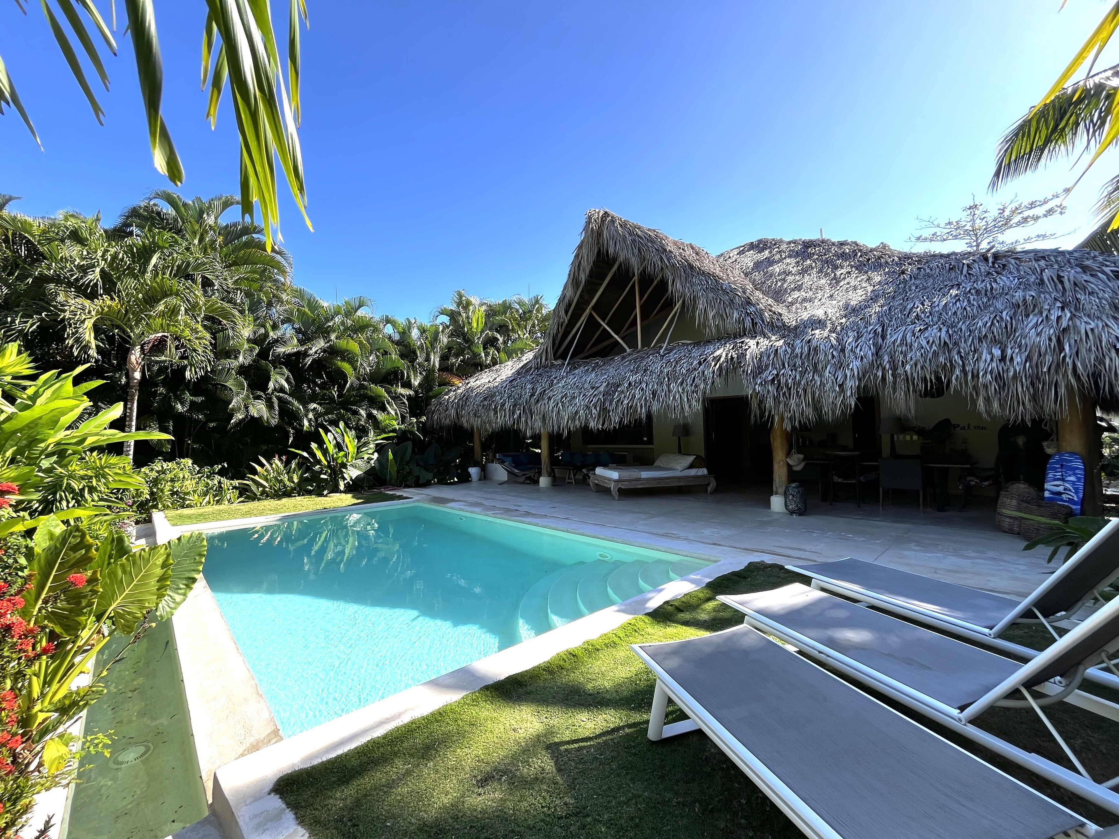Zona de piscina con una estructura de techo de paja, sillas de descanso y abundante vegetación tropical.