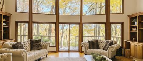 A spacious living room with large windows offering a view of autumn trees. There are two sofas and a coffee table with decorative plants.