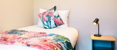 Una cama bien hecha con un edredón colorido y una almohada decorativa, una mesita de noche con una lámpara y un estante azul.