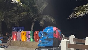 A public space with a large skull sculpture, colorful letters spelling "MORE," and palm trees.
