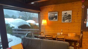 Ein gemütlicher Raum mit Holzvertäfelung, einer Couch mit Kissen, einem Esstisch mit Stühlen und einem Fenster mit Blick auf eine verschneite Landschaft.