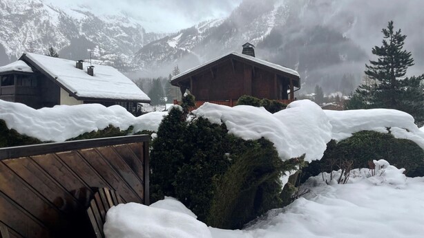 Eine Holzhütte mit geneigtem Dach, umgeben von schneebedeckten Bäumen und Bergen im Hintergrund.