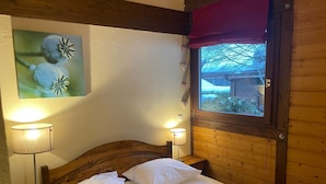 Ein holzgetäfeltes Zimmer im Blockhausstil mit einem Bett, zwei Nachttischlampen, einem Fenster mit Blick auf eine verschneite Landschaft und einem an der Wand hängenden Bild von Blumen.