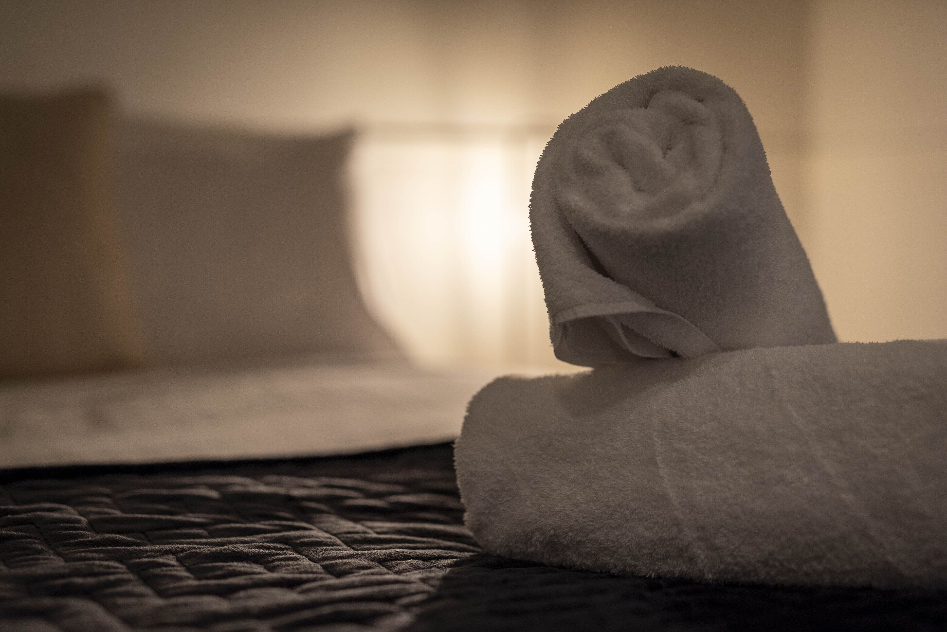 Une serviette blanche, soigneusement pliée, est posée sur un lit recouvert d’une couverture à motifs de couleur sombre.
