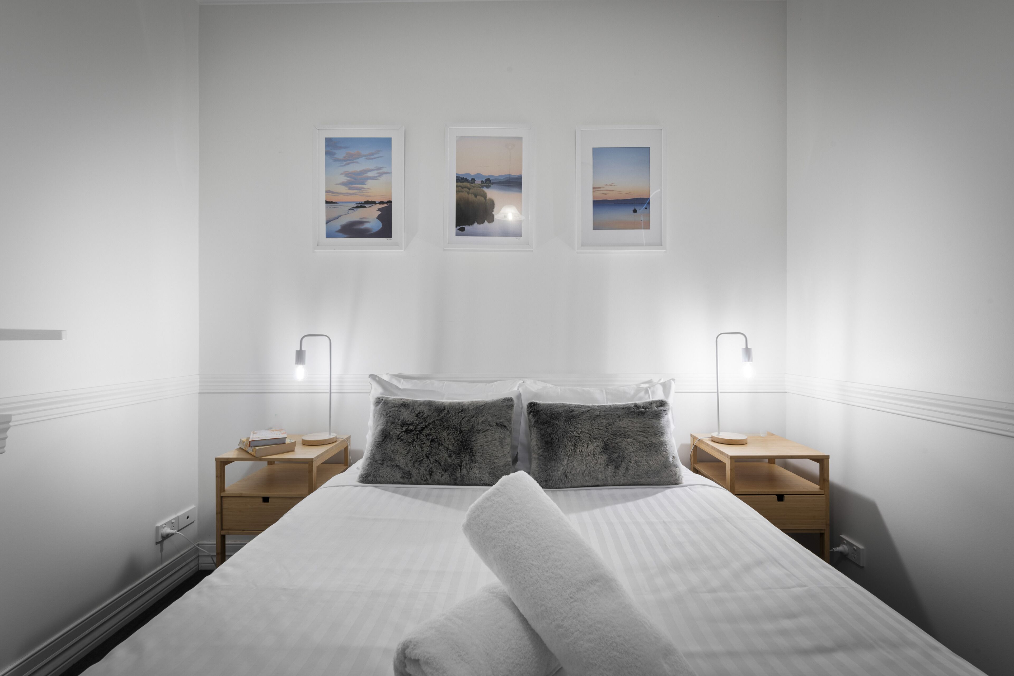 Une chambre bien rangée, avec un lit, deux tables de chevet et trois tableaux encadrés accrochés au mur.