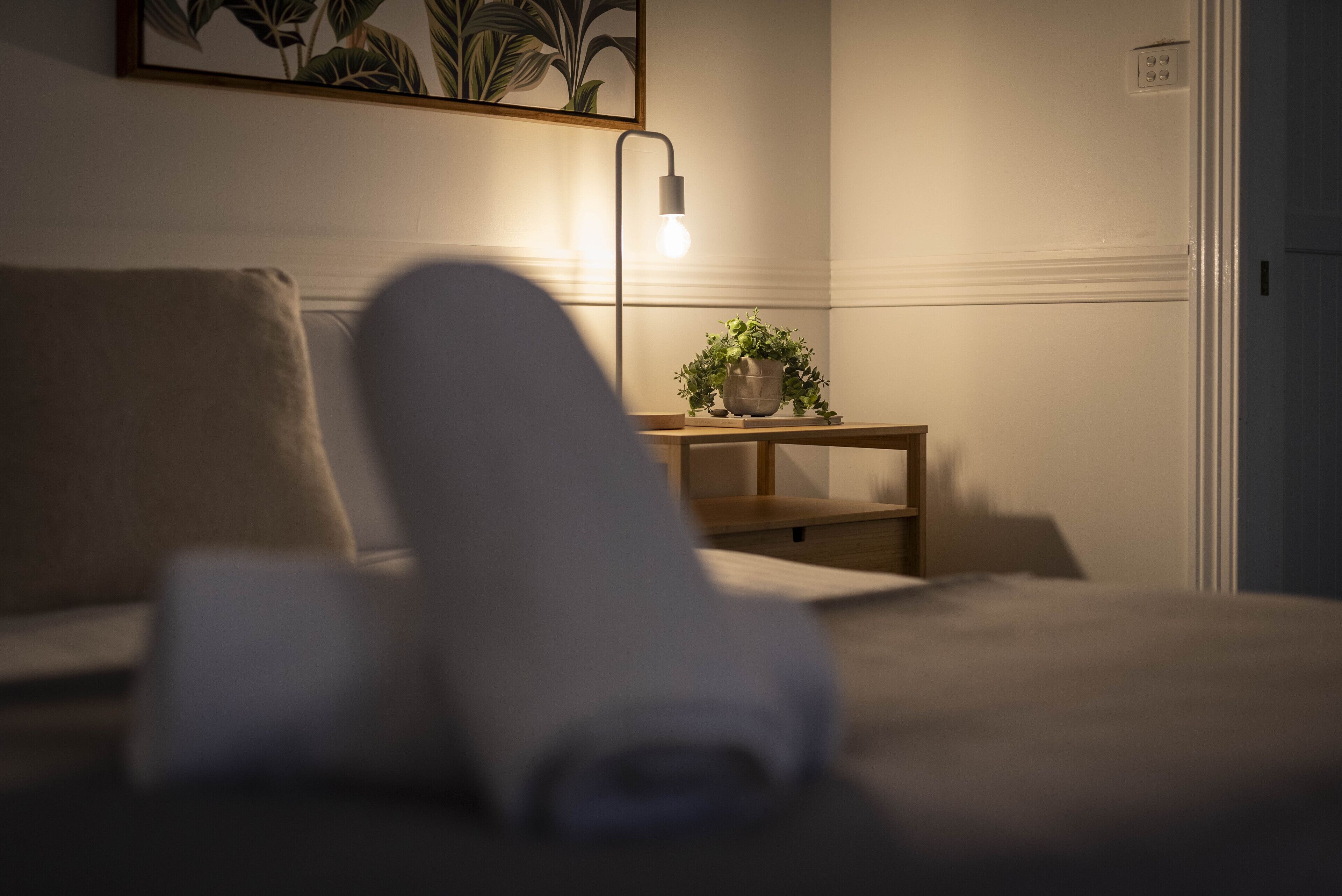 Una cama bien hecha con una toalla enrollada, una mesita de noche con una planta y una lámpara fijada a la pared.