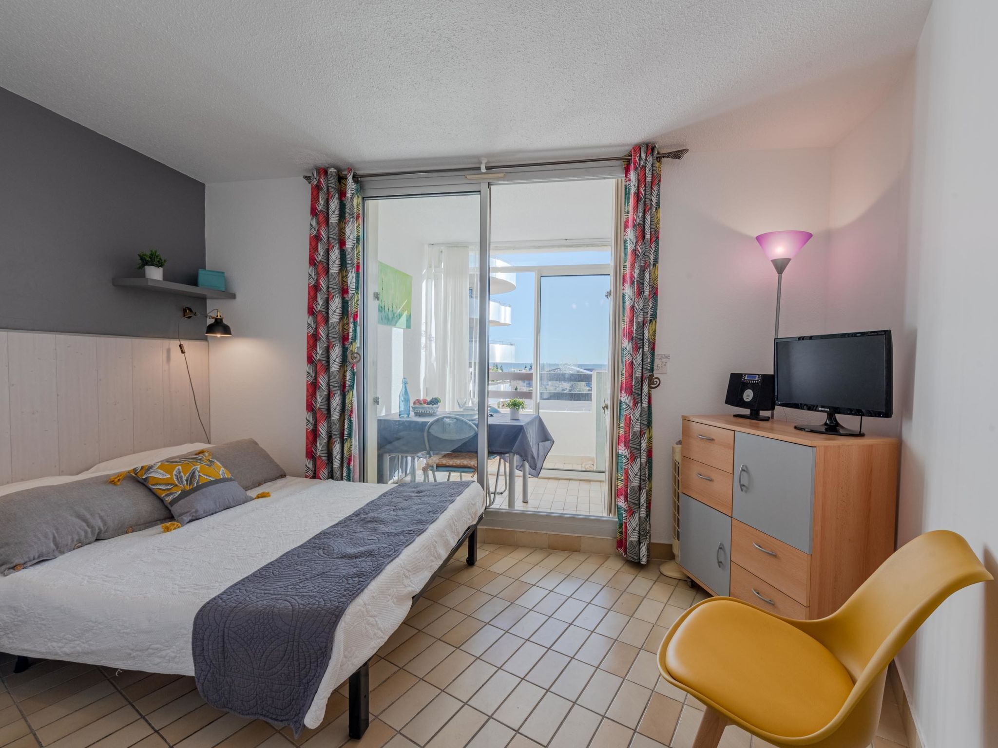 A bedroom with a bed, a desk, a chair, a TV, and a balcony with curtains.