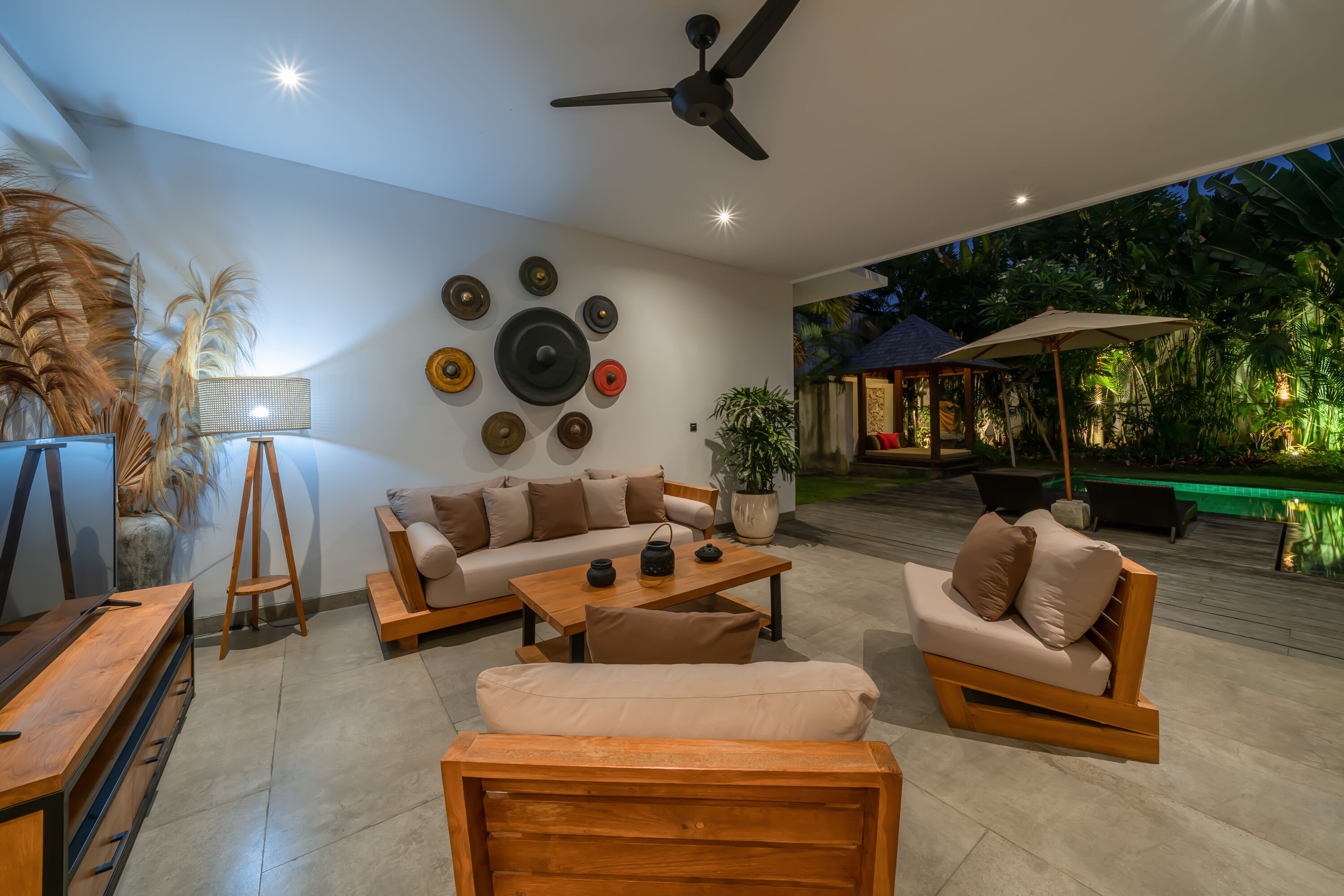 Un salon moderne avec un canapé, une table basse et des chaises en bois. On y trouve un ventilateur de plafond, un luminaire fixé au mur et une plante en pot.