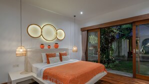 A modern bedroom with a large bed, a nightstand, a wall-mounted light fixture, and a sliding glass door leading to a garden. - Villa Lisy  (Canggu)