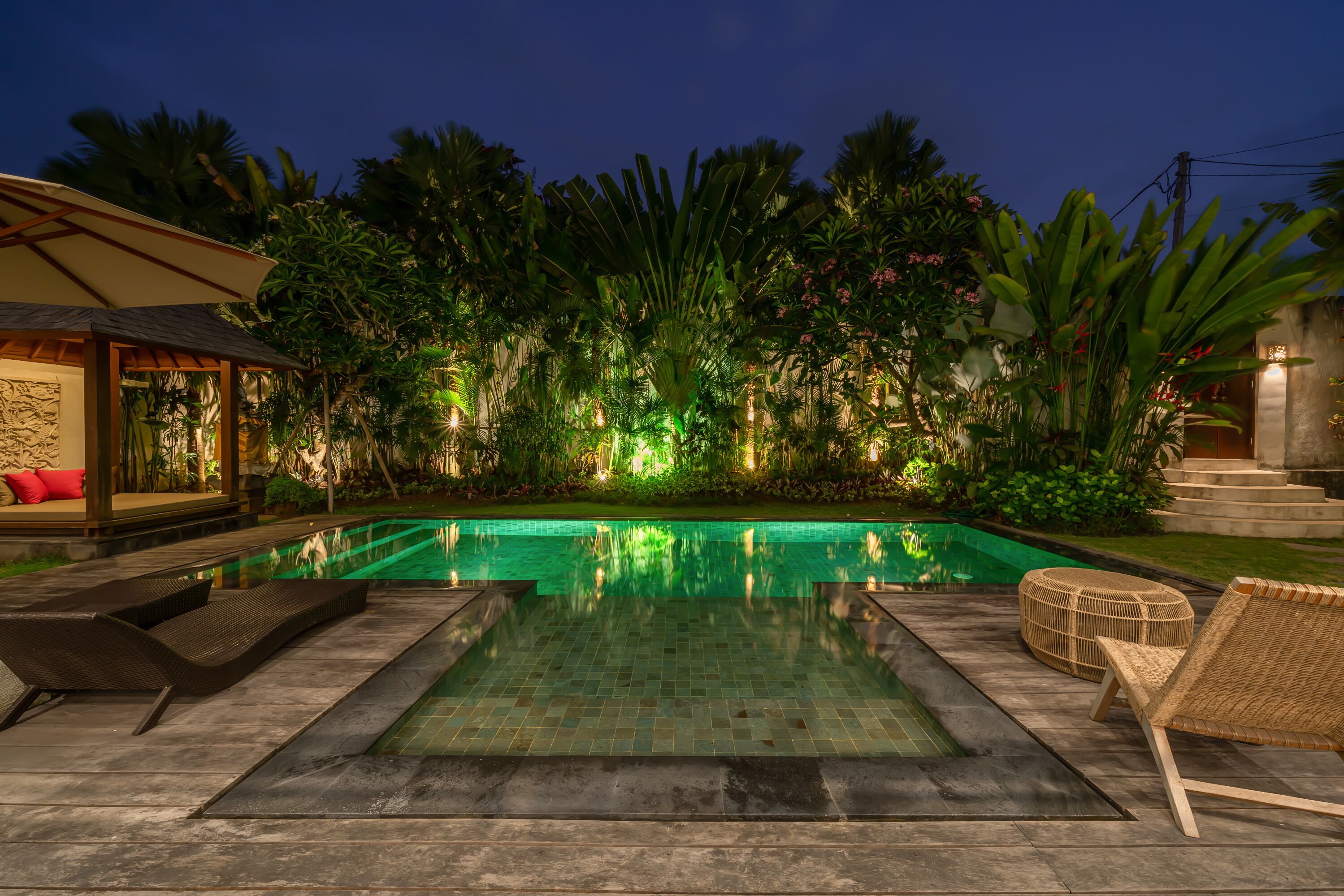 A swimming pool with lounge chairs, surrounded by lush greenery and a well-lit patio area.