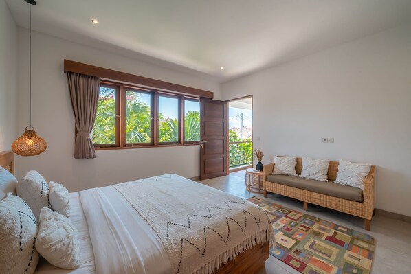 A bedroom with a large bed, a sofa, a window with curtains, and a view of greenery outside. - Villa Lisy (Canggu)