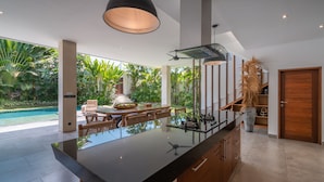 A modern kitchen with a large island, a dining area with wicker furniture, and a view of a pool and lush greenery. - Villa Lisy  (Canggu)