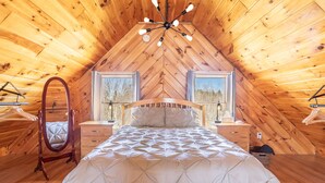 Ein gemütliches Schlafzimmer im Dachgeschoss mit Holzbalkendecke, einem Bett mit gesteppter Bettdecke, zwei Nachttischen mit Lampen und einem Hängestuhl.
