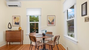 Un coin repas avec une table ronde, six chaises et un buffet en bois. Il y a deux fenêtres avec des stores blancs, et des œuvres d’art sont exposées sur les murs.
