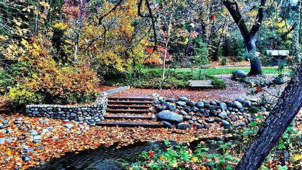 Un pont de pierre enjambant un ruisseau, bordé de feuilles d’automne, entouré d’arbres et d’un mur de pierres.