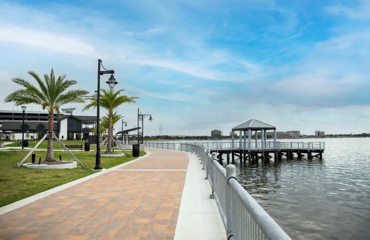 A waterfront walkway with a gazebo, palm trees, and a dock extending into the water.