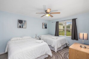 A bedroom with two beds, a ceiling fan, a window with curtains, and a bedside table with a lamp.