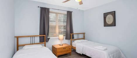 A bedroom with two single beds, a ceiling fan, a window with curtains, and a bedside table with a lamp.