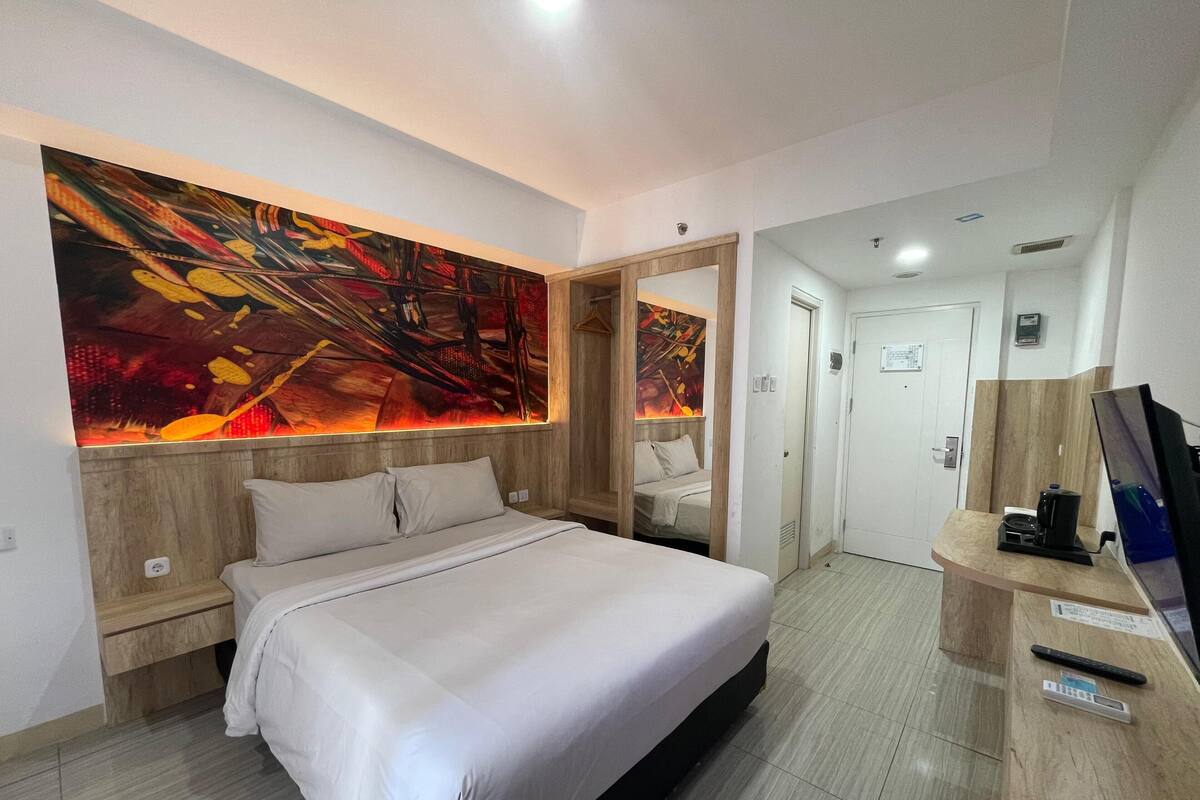 A modern hotel room with a large bed, a wooden headboard, a large abstract painting, a mirror, a desk with a coffee maker, and a TV.