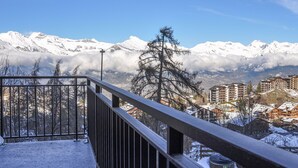 Ein Balkon mit Metallgeländer mit Blick auf eine verschneite Berglandschaft, im Hintergrund sind Gebäude zu sehen.