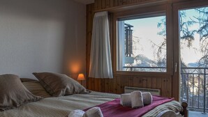 Ein Schlafzimmer mit großem Fenster, einem Bett mit karierter Bettwäsche und Blick auf Bäume und ein Gebäude.