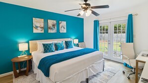 A bedroom with a bed, a desk, a chair, a ceiling fan, and a view of the outdoors.