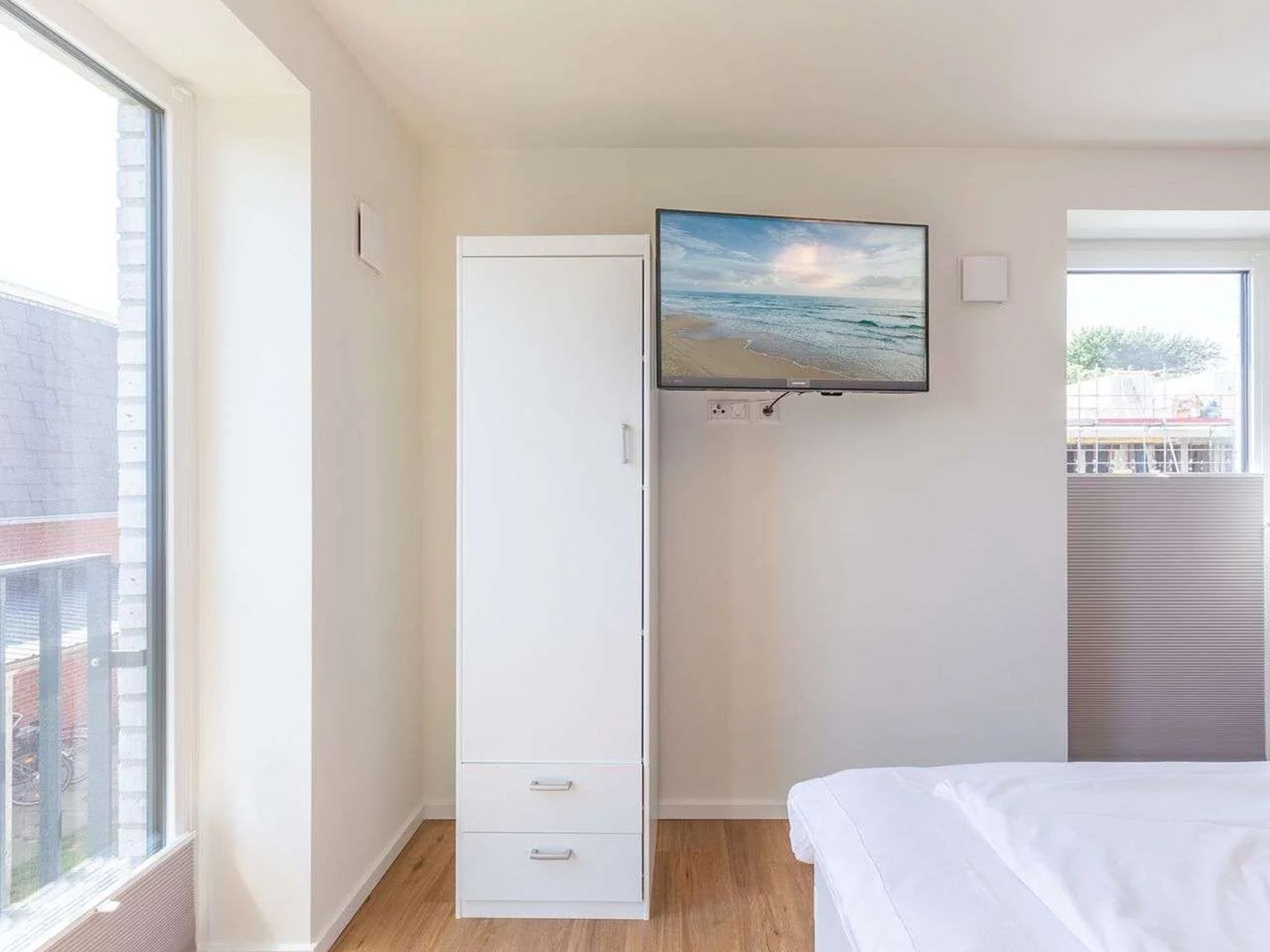 A bedroom with a bed, a white wardrobe, a television mounted on the wall, and a window with a view of the outside.