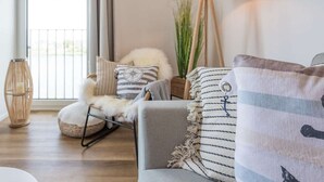 Ein gemütliches Wohnzimmer mit einem grauen Sofa, gestreiftem Kissen, einem flauschigen Sessel, einer Stehlampe und Blick durch eine Schiebetür.