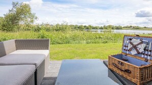 A patio with a wicker sofa and a picnic basket, overlooking a lake and grassy area. - Liselotte by Interhome (Nordstrand)