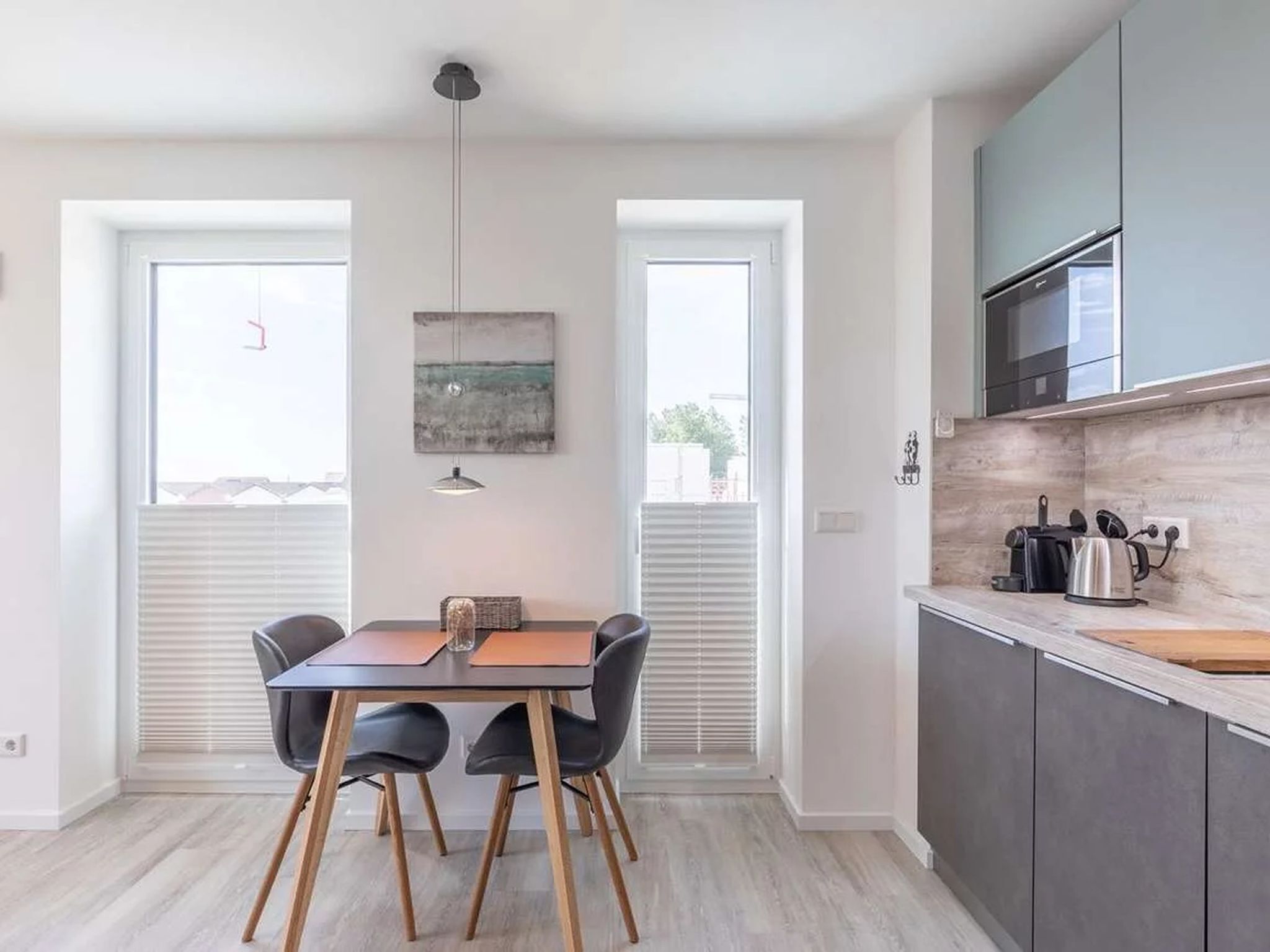 A modern kitchen with grey cabinets, a built-in oven, and a countertop with a kettle and other appliances. There is a dining area with a table and chairs, and a window with blinds.