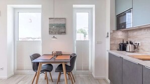A modern kitchen with grey cabinets, a built-in oven, and a countertop with a kettle and other appliances. There is a dining area with a table and chairs, and a window with blinds. - Heewer Stuuv by Interhome (Nordstrand)
