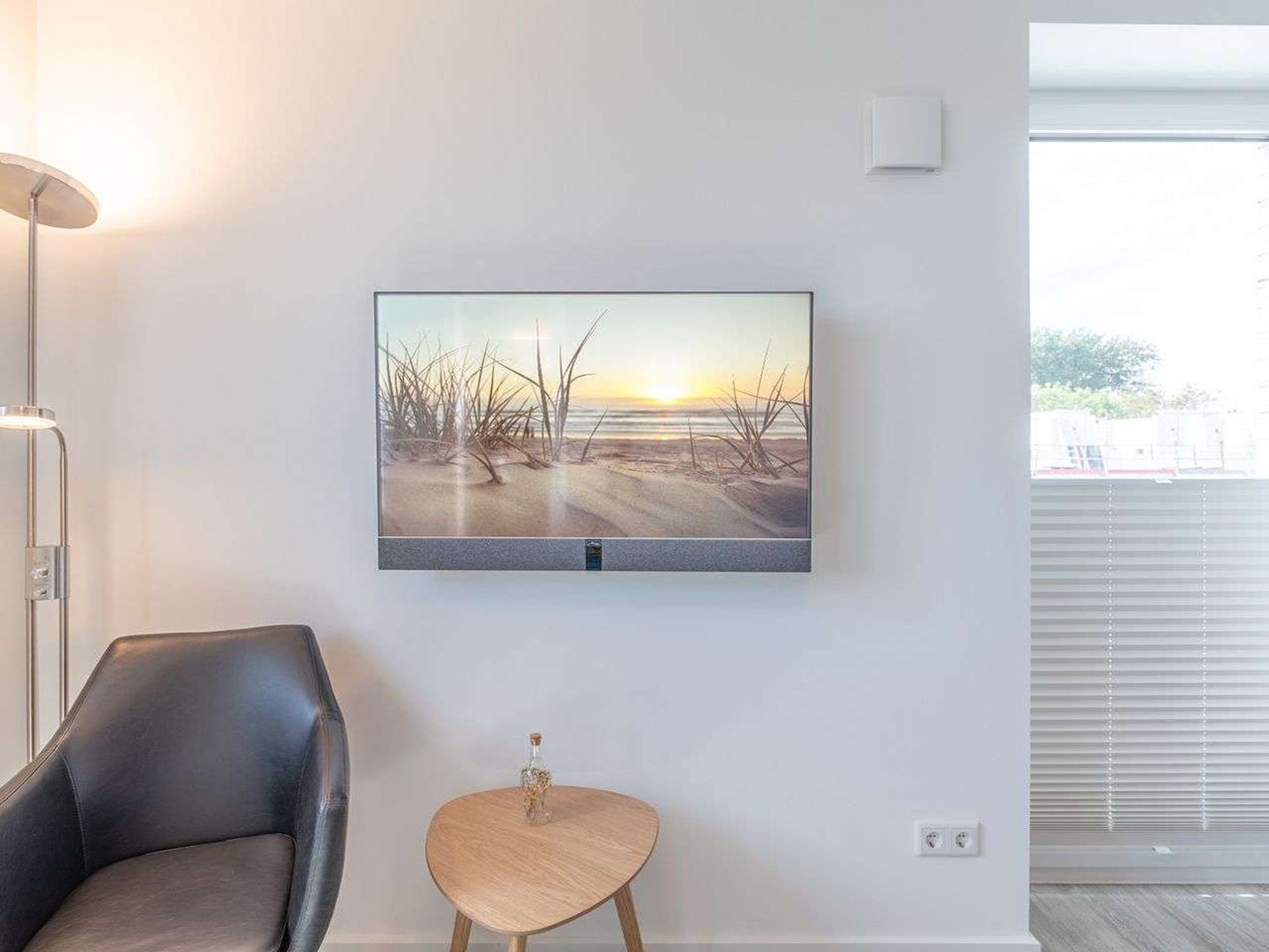 A modern room with a large flat-screen TV mounted on the wall, a leather chair, a small wooden side table, and a window with blinds.