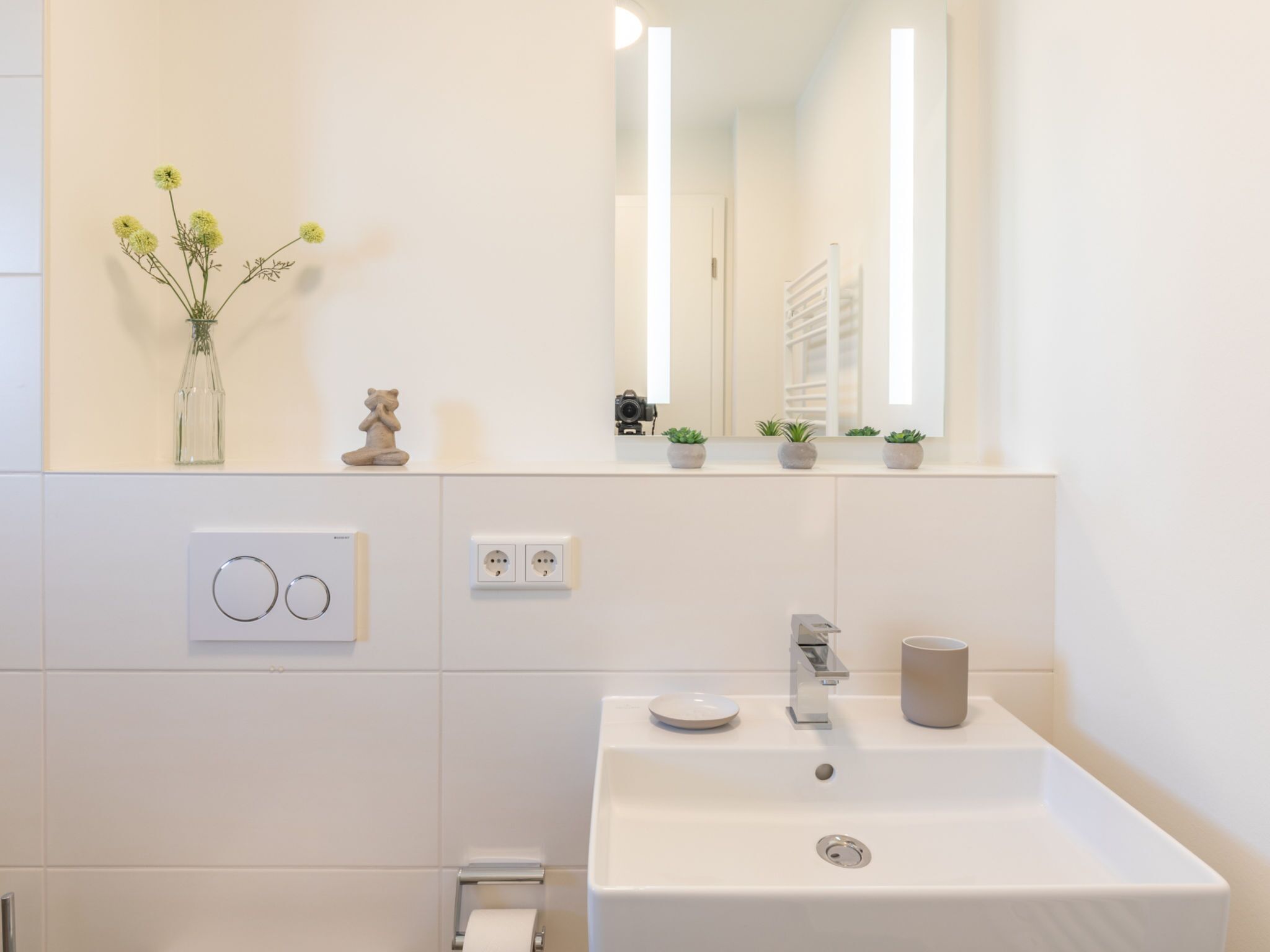 Une salle de bain moderne avec un lavabo blanc, un miroir et un vase rempli de fleurs jaunes.