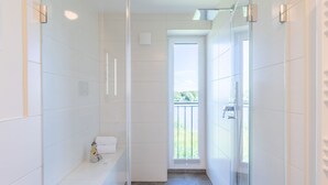 A modern bathroom with a glass shower enclosure, a bench with towels, and a view of greenery outside. - Die Komfortzone by Interhome (Nordstrand)