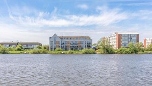 Blick auf einen modernen Apartmentkomplex an einem Flussufer, im Vordergrund ein ruhiger Fluss und ein klarer Himmel.